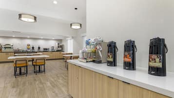 A modern kitchen area with a white countertop, coffee machines, and a seating area with wooden chairs.