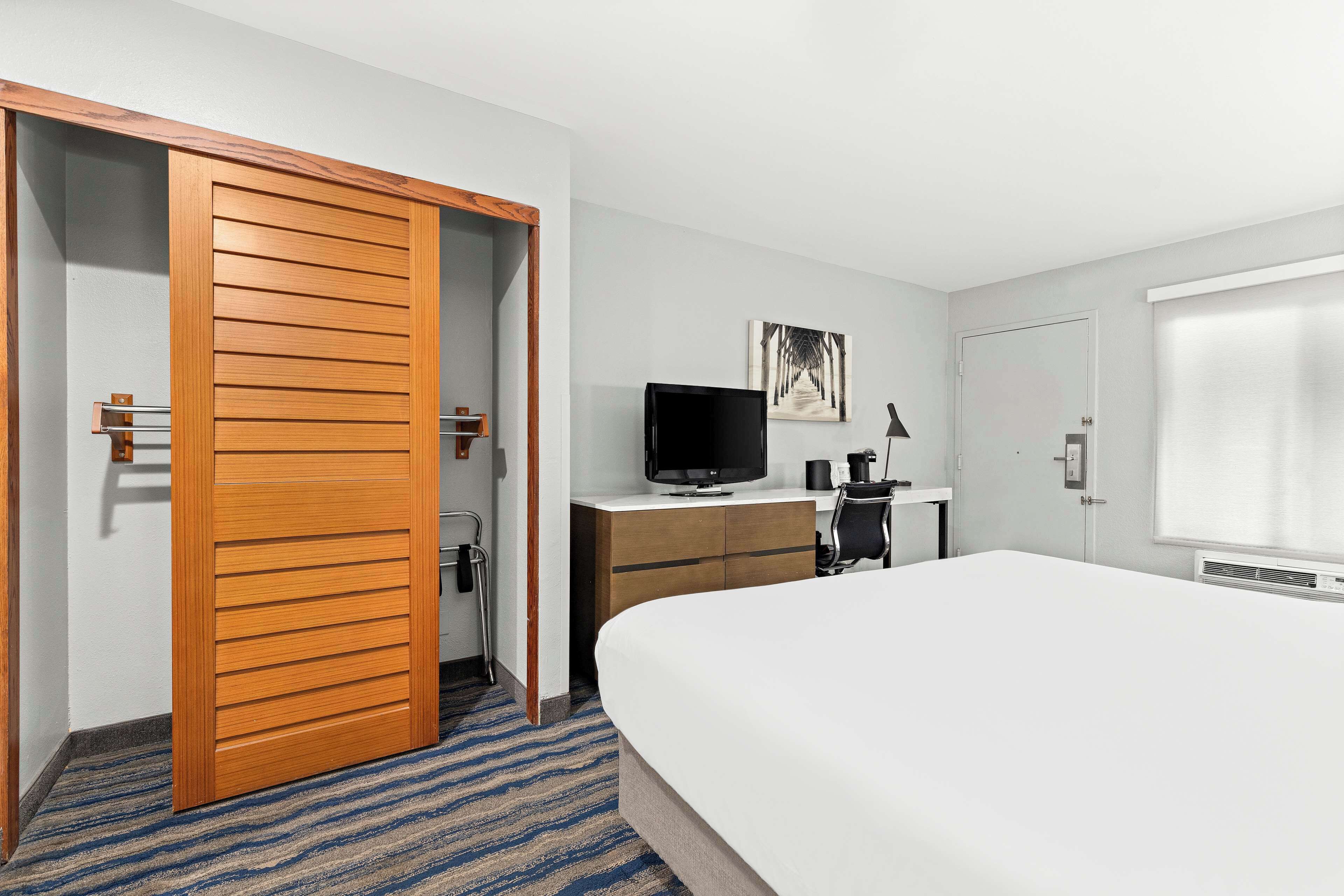 A hotel room with a large bed, a wooden wardrobe, a desk with a television, and a painting on the wall.