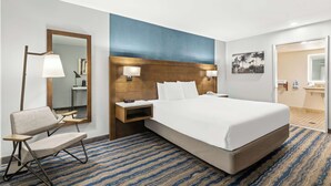 A hotel room with a large bed, a desk with a chair, a nightstand, a mirror, and a blue-striped carpet.