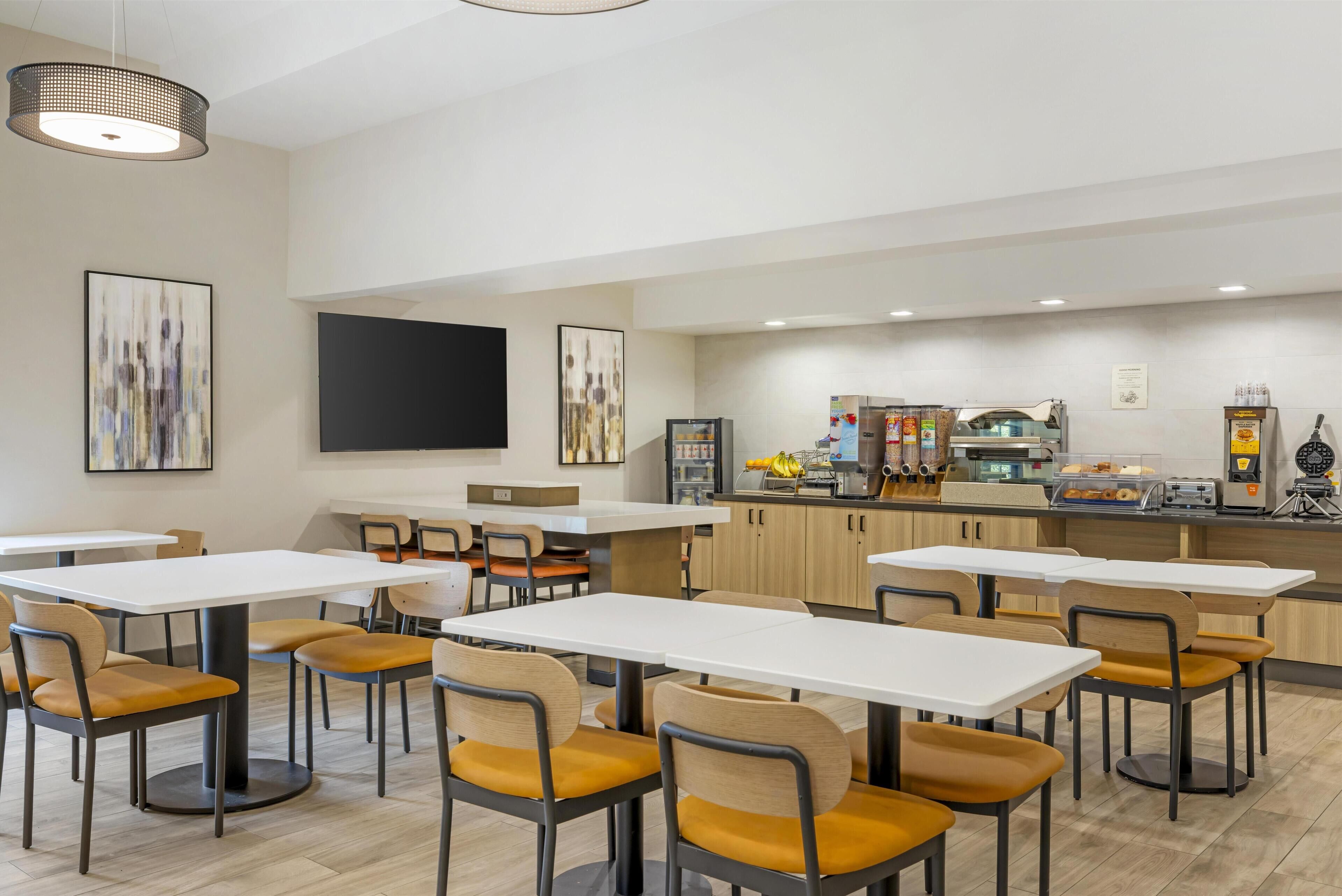 A modern dining area with white tables and chairs, a kitchen counter with appliances, and artwork on the walls.