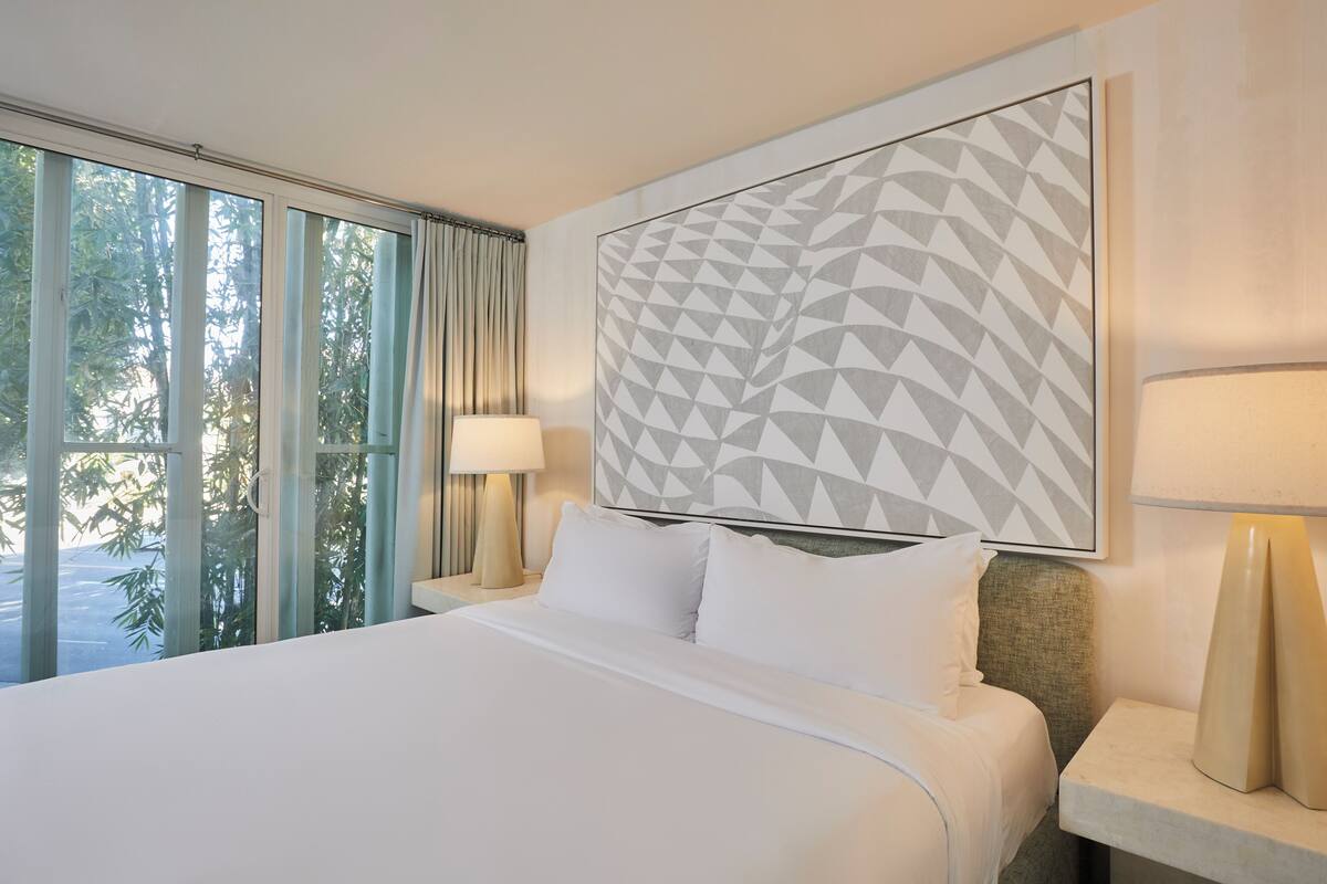 Una habitación de hotel con una cama grande, una mesita de noche con lámpara, una ventana con cortinas y un cuadro con diseño geométrico.