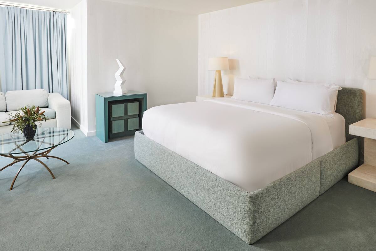 Habitación de hotel moderna con una cama grande, lámparas de noche, una mesita auxiliar pequeña, una mesa de centro de vidrio y vistas al exterior.