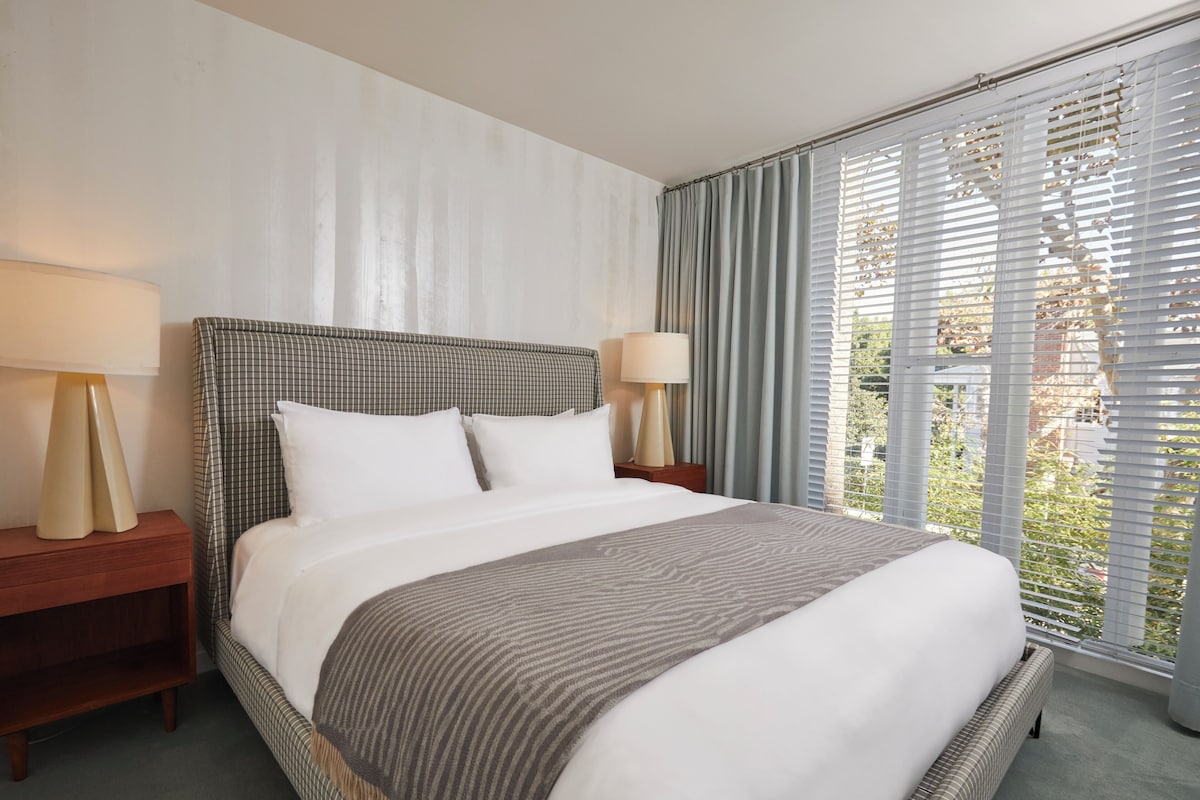 Een net opgemaakt bed met een geruit hoofdbord, twee nachtlampjes en uitzicht door een raam met jaloezieën.