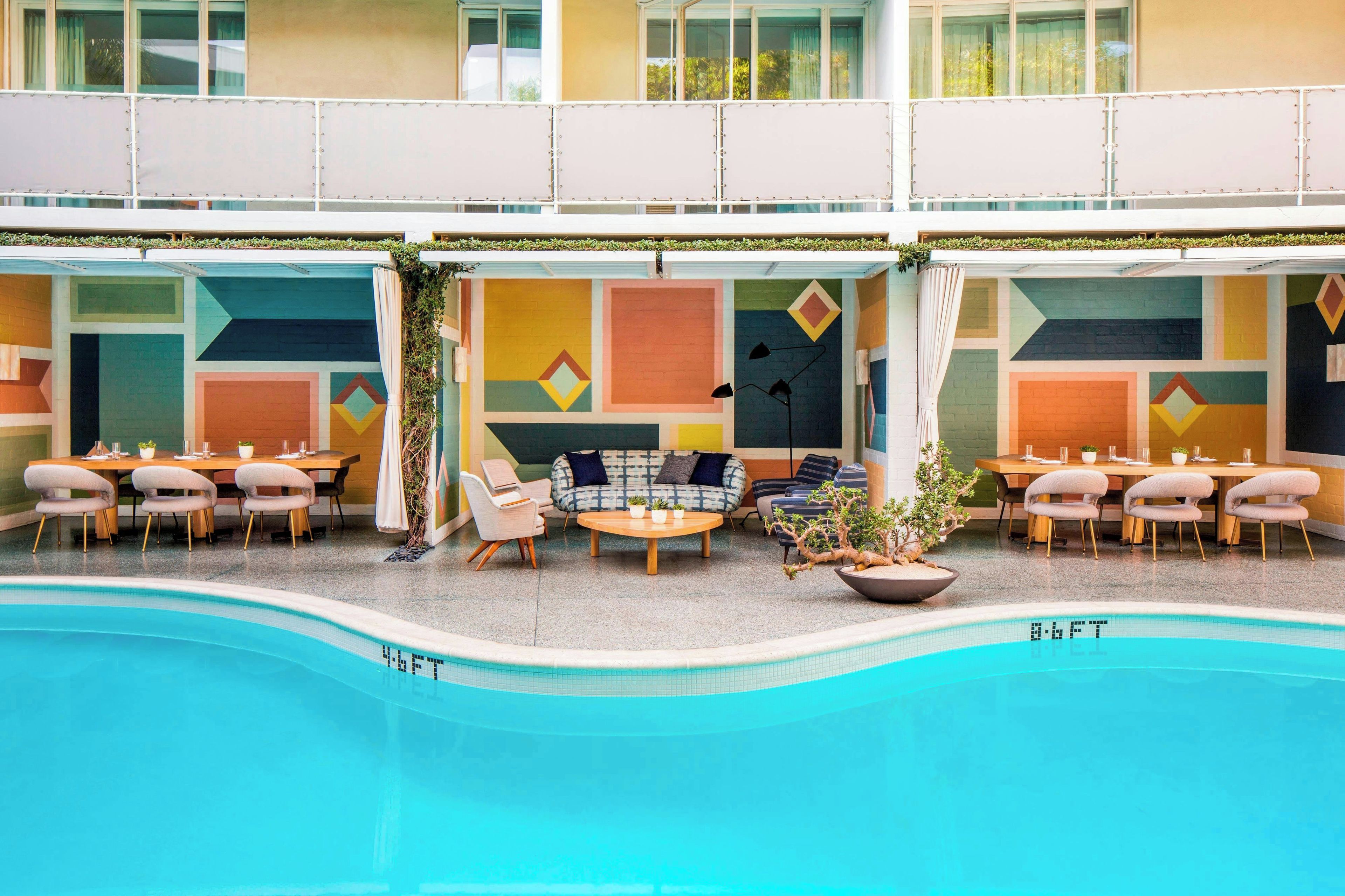 Zona junto a la piscina con mobiliario exterior, una piscina y un edificio con arte mural geométrico de colores.