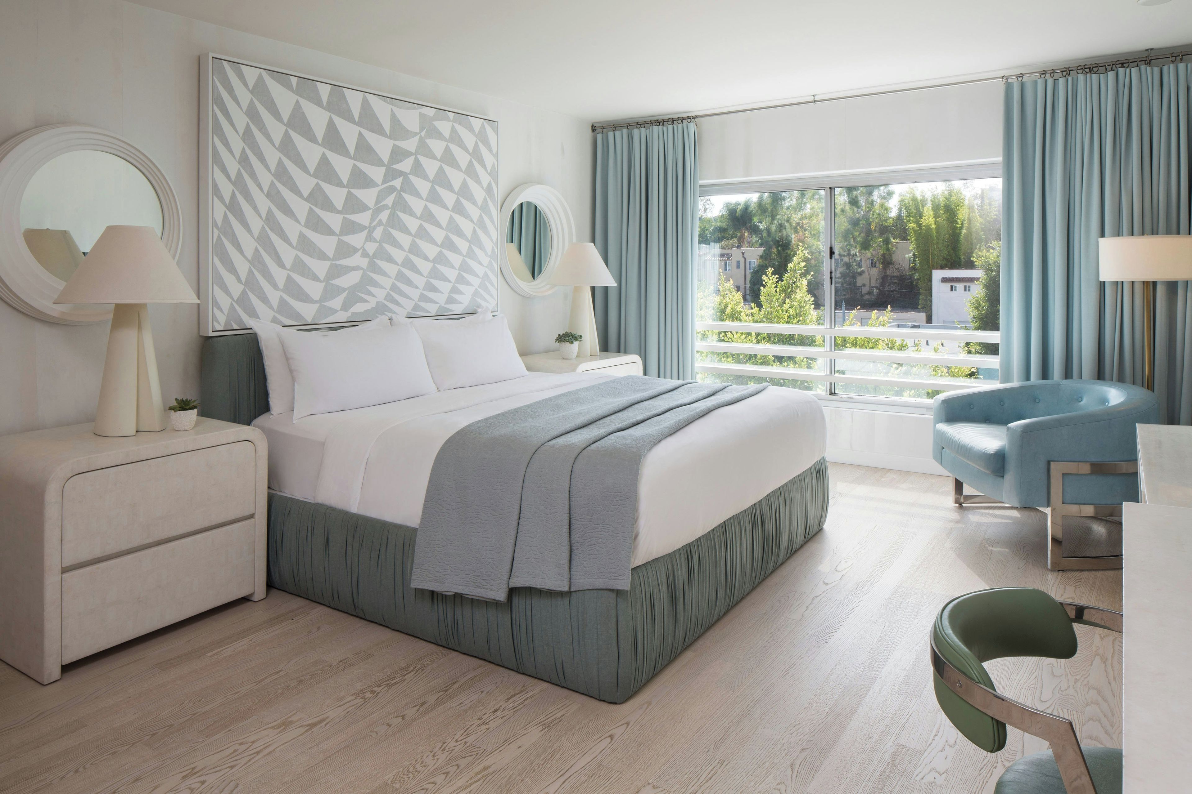 A modern bedroom with a large bed, bedside lamps, a chair, and a view of greenery outside.