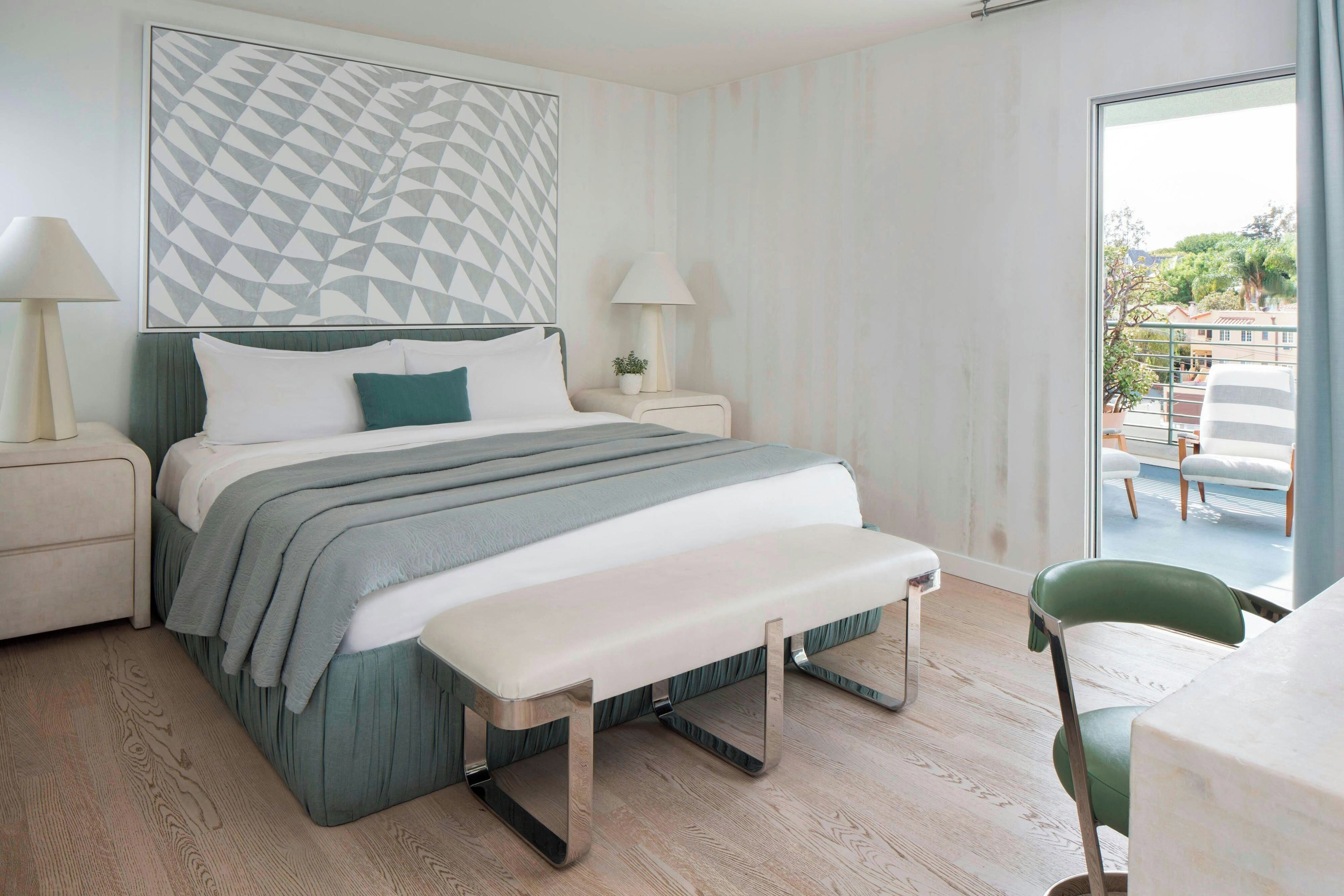 Quarto de hotel com cama, mesas de cabeceira, um banco e vista para o exterior, além de uma obra de arte geométrica na parede.