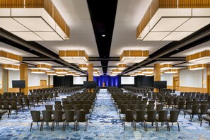 Un salón de conferencias con filas de sillas, una pantalla grande y un telón de fondo azul.