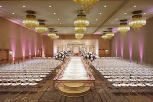 Un amplio salón de banquetes con filas de mesas y sillas, un escenario central y candelabros ornamentales.