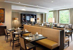 A hotel dining area with a long bench, multiple tables, and chairs, a television, and a large window with a view of trees. - Hyatt Regency Bellevue (Bellevue)