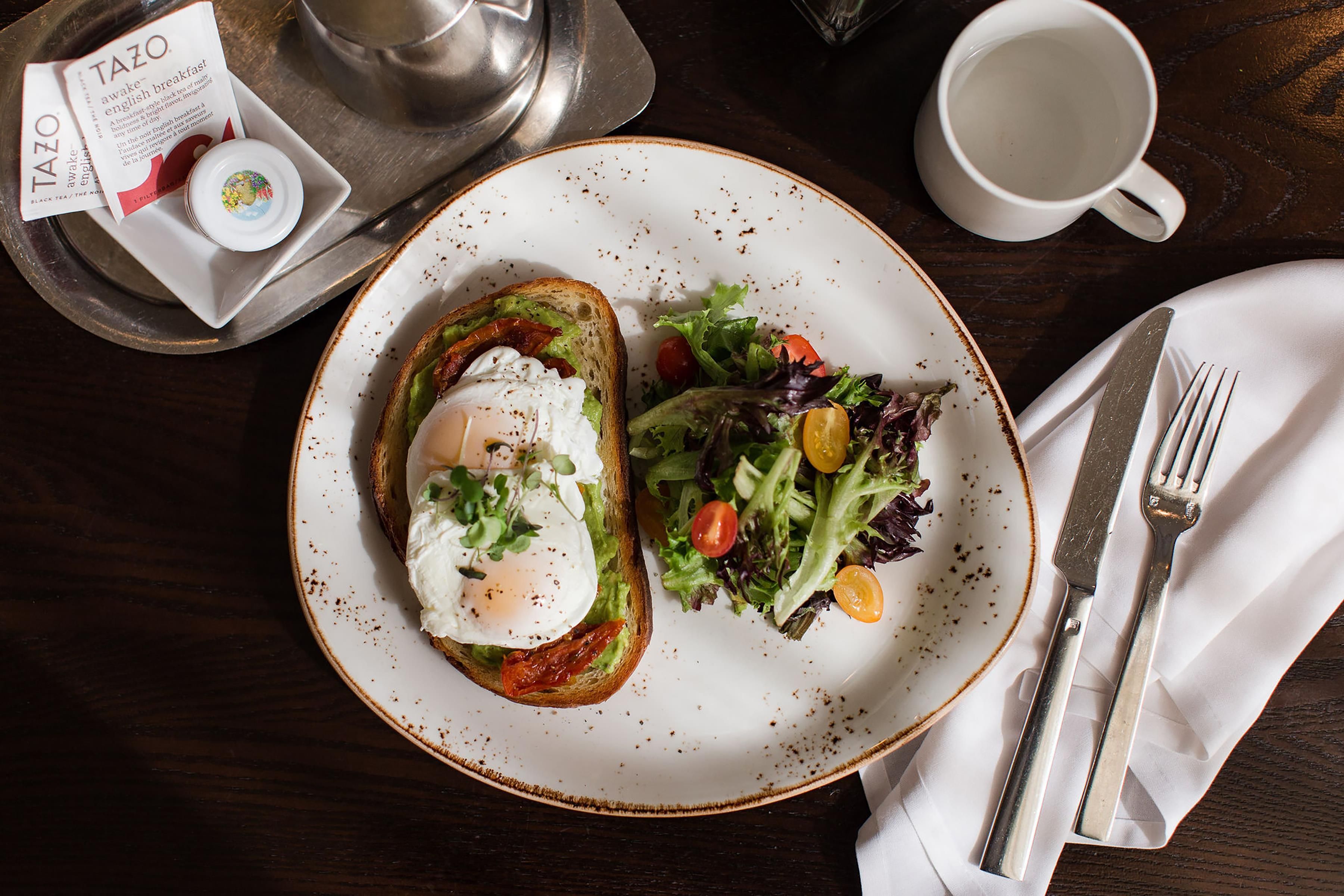 A plate with toast topped with avocado, a poached egg, and greens, a cup of coffee, and a menu on a tray.