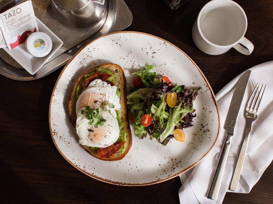 A plate with toast topped with avocado, a poached egg, and greens, a cup of coffee, and a menu on a tray.