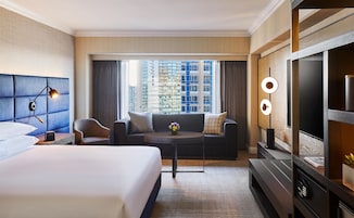 Quarto de hotel com cama, sofá, mesa de centro, televisão e vista para o horizonte urbano através de amplas janelas.