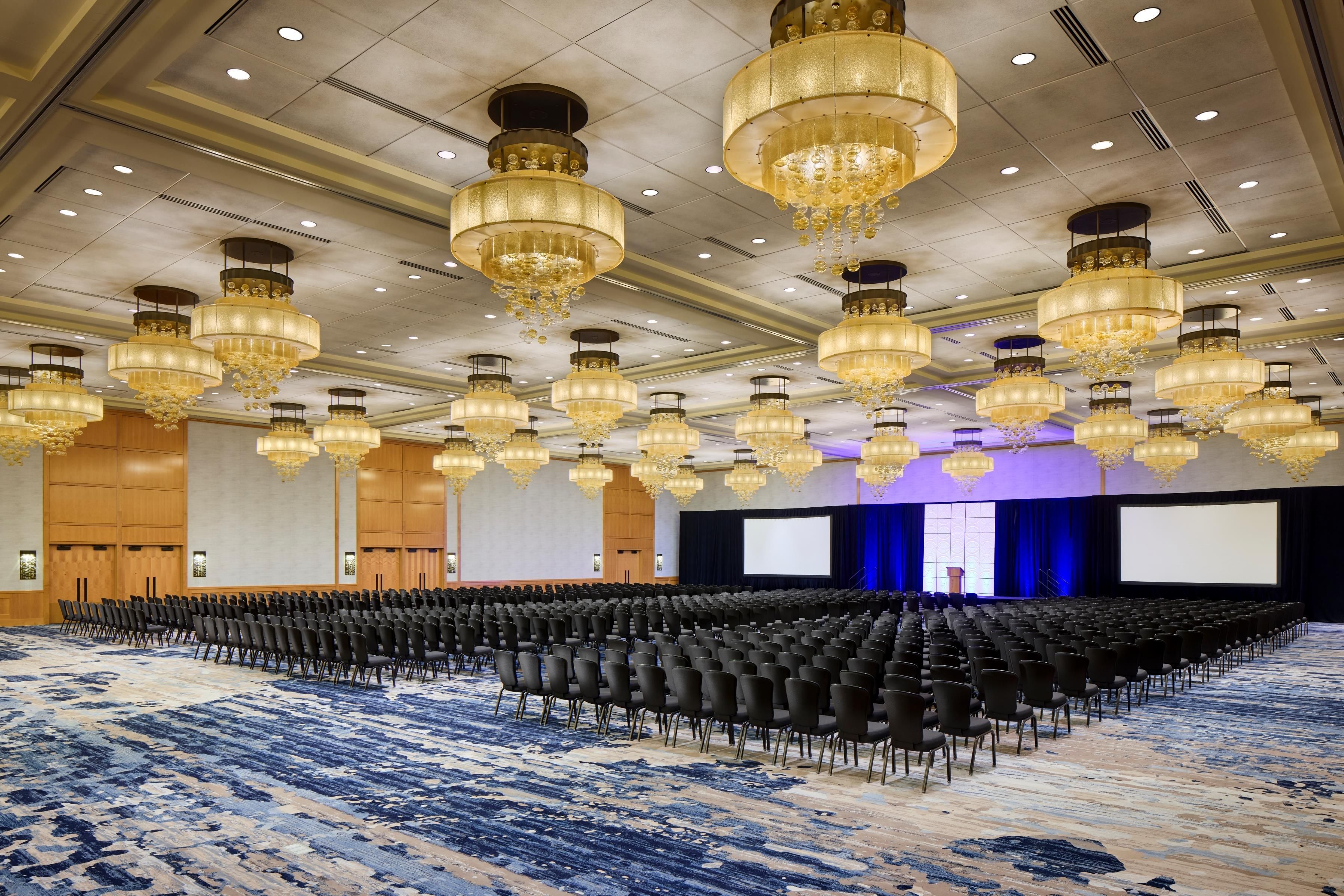A large conference room with rows of chairs, a stage with a screen, and multiple chandeliers.