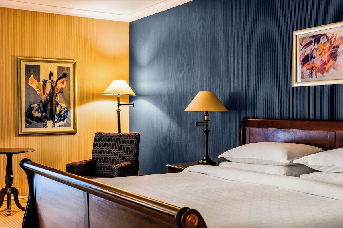 Une chambre d’hôtel avec un lit, deux lampes, une chaise, une petite table et un tableau au mur.