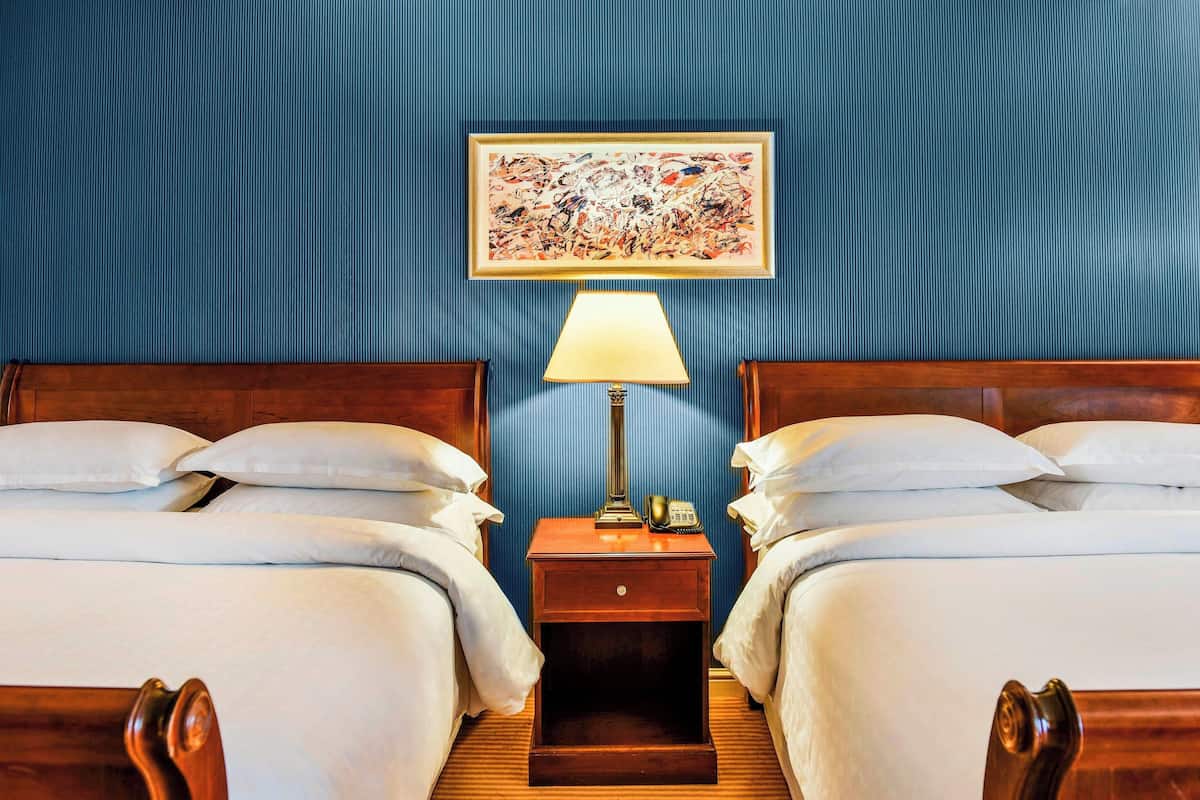 Une chambre d’hôtel avec deux lits, une table de chevet avec une lampe, un téléphone et un tableau au mur.