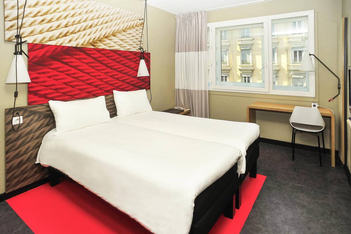 Hotelkamer met een groot bed, een bureau, een stoel, een raam met uitzicht op gebouwen en een rood tapijt.