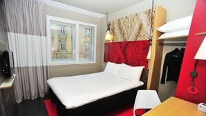 A hotel room with a bed, a desk, a chair, a TV, a window with curtains, and a view of buildings outside. - Ibis Lyon Centre (Lyon)
