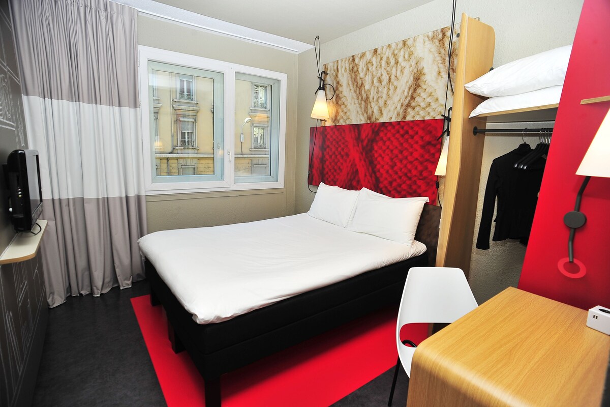A hotel room with a bed, a desk, a chair, a TV, a window with curtains, and a view of buildings outside.