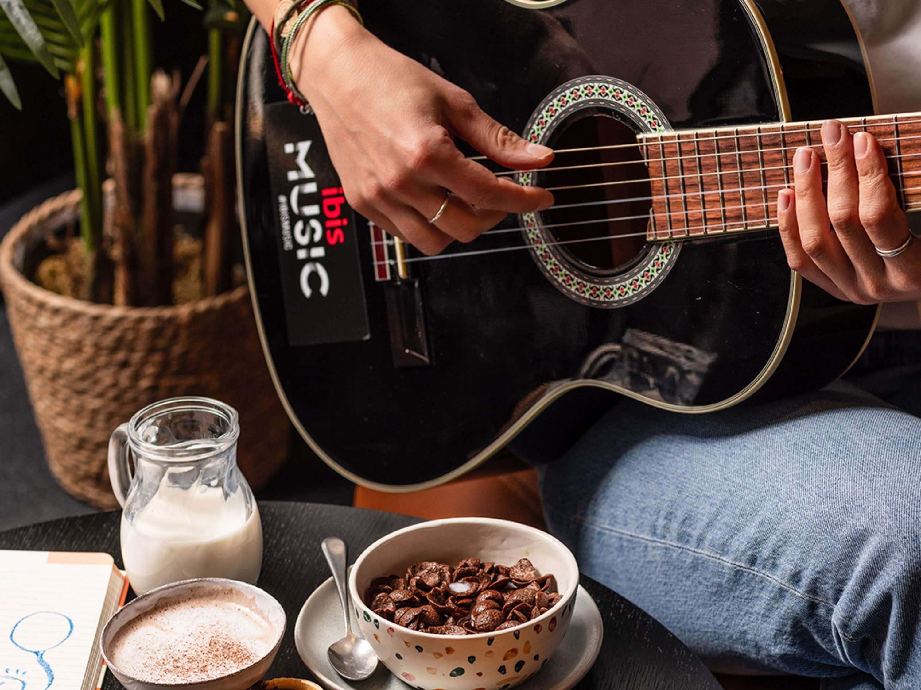 A person playing a black guitar with a 'MUSIC' label, seated next to a table with a milk jug, a bowl of cereal, and a cup of coffee.