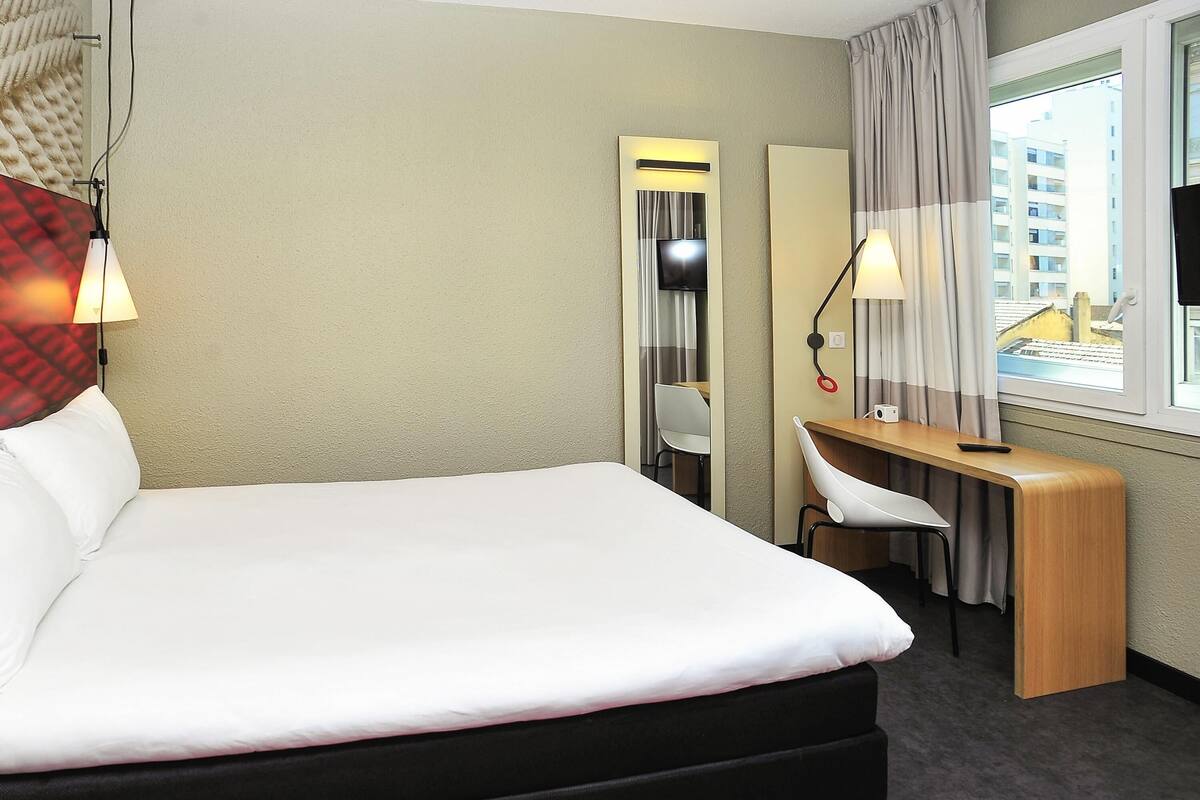 A hotel room with a bed, desk, chair, and a window with a view of buildings.