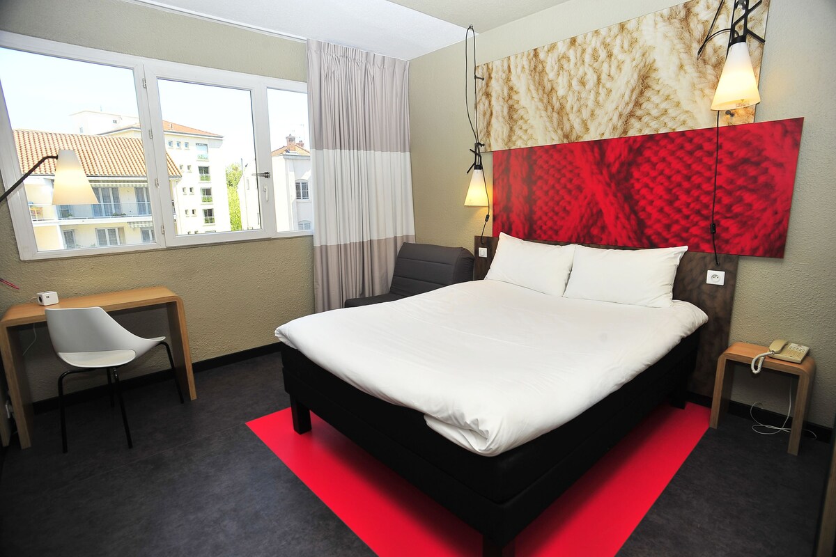 A hotel room with a large bed, a desk, a chair, a telephone, and a window with curtains.