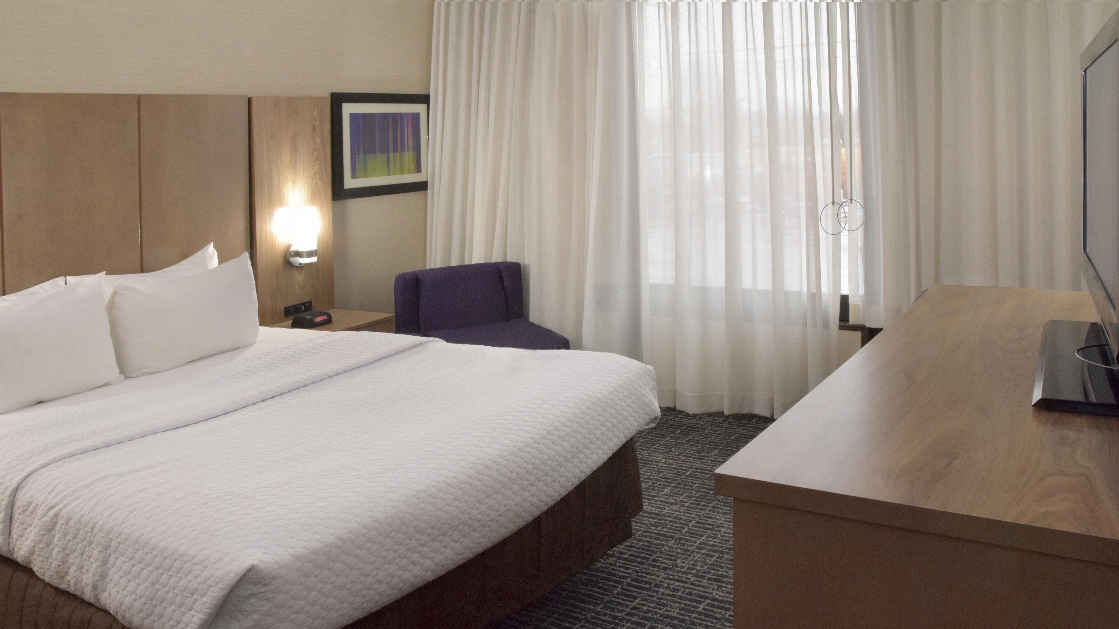 Een hotelkamer met een groot bed, een bureau, een stoel, een nachtkastje en een raam met dunne gordijnen.