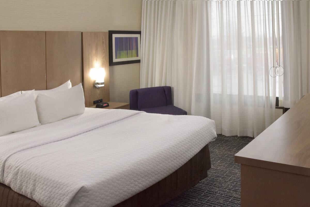 Een hotelkamer met een groot bed, een bureau, een stoel, een nachtkastje, een lamp en een raam met gordijnen.