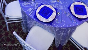 Een ronde tafel met een blauwe tafelkleed, witte stoelen en blauwe middelpuntdecoraties.