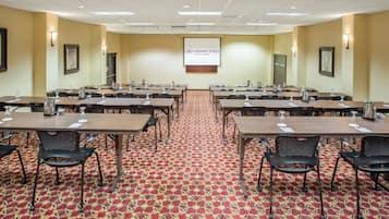Un salón de conferencias con varias mesas y sillas, piso alfombrado y una pantalla de proyección en la pared.