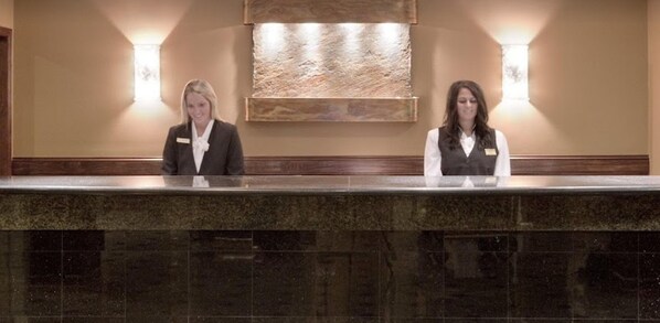 A reception desk with two staff members, a stone wall with a textured artwork, and wall-mounted lighting. - Crowne Plaza Cleveland Airport by IHG (Middleburg Heights)