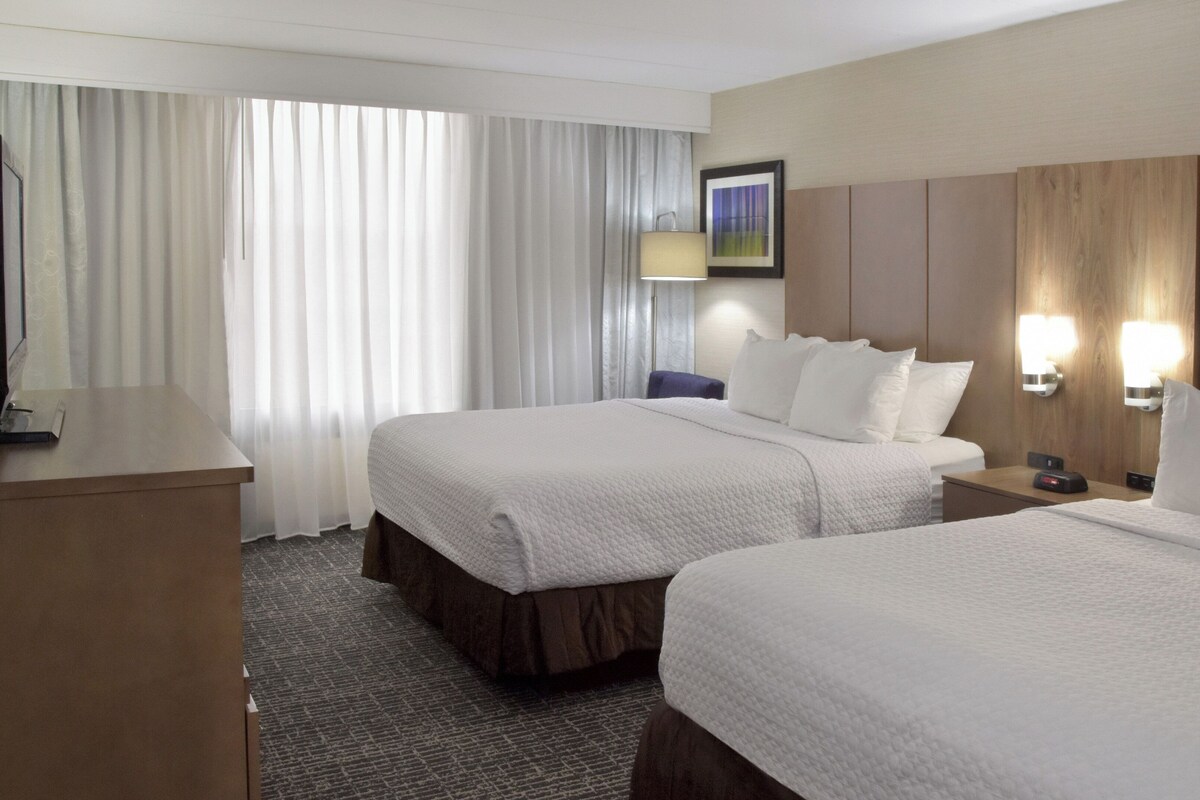 A hotel room with two beds, a desk, a chair, a TV, and a painting on the wall.