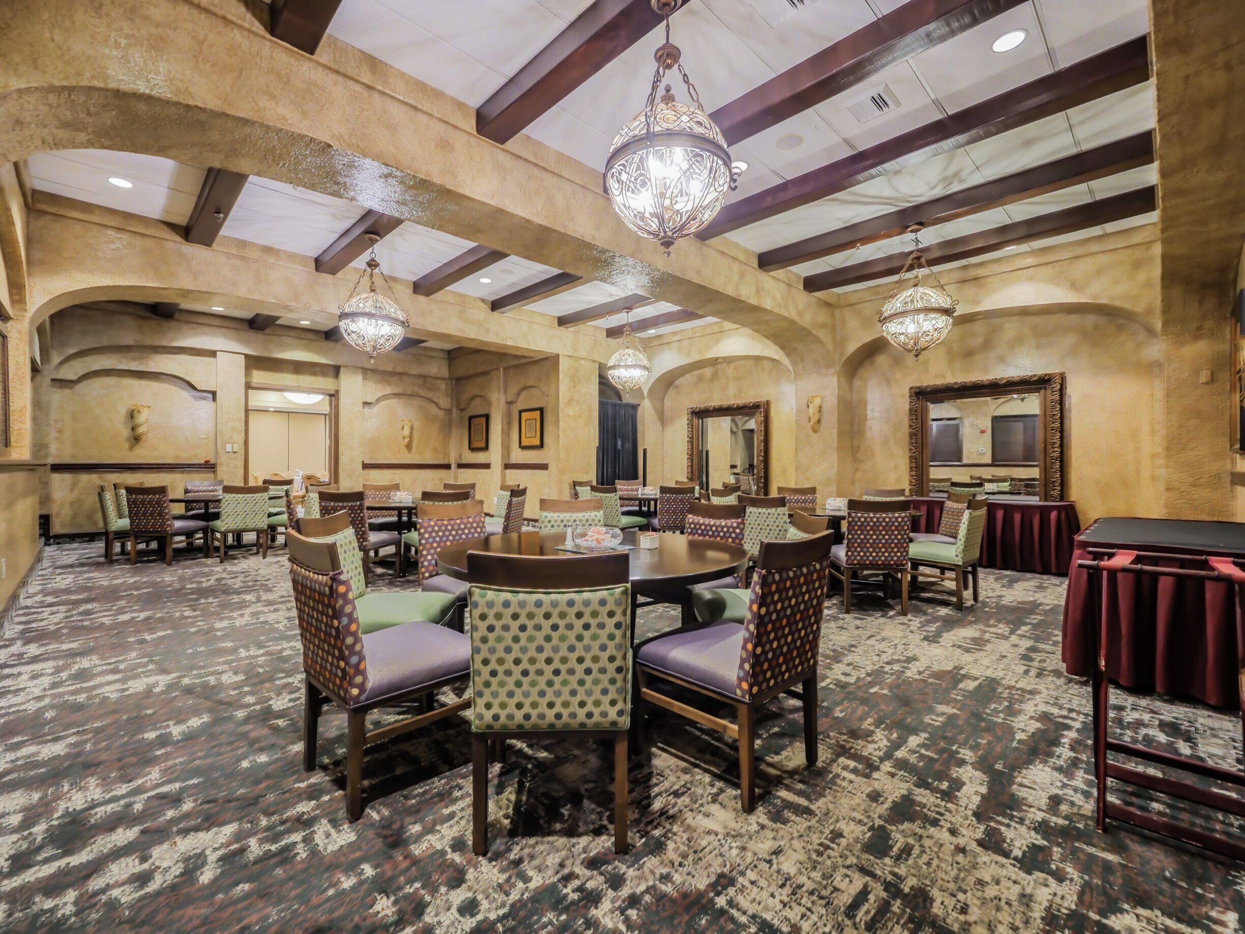 A hotel dining area with multiple tables and chairs, chandeliers, and a patterned carpet.