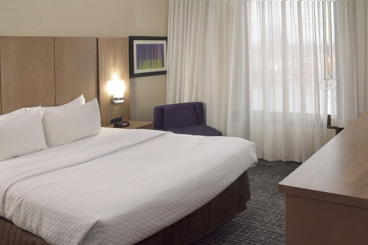 A hotel room with a bed, a desk, a chair, a nightstand, a lamp, and a window with curtains.