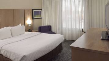 A hotel room with a bed, a desk, a chair, a nightstand, a lamp, and a window with curtains.
