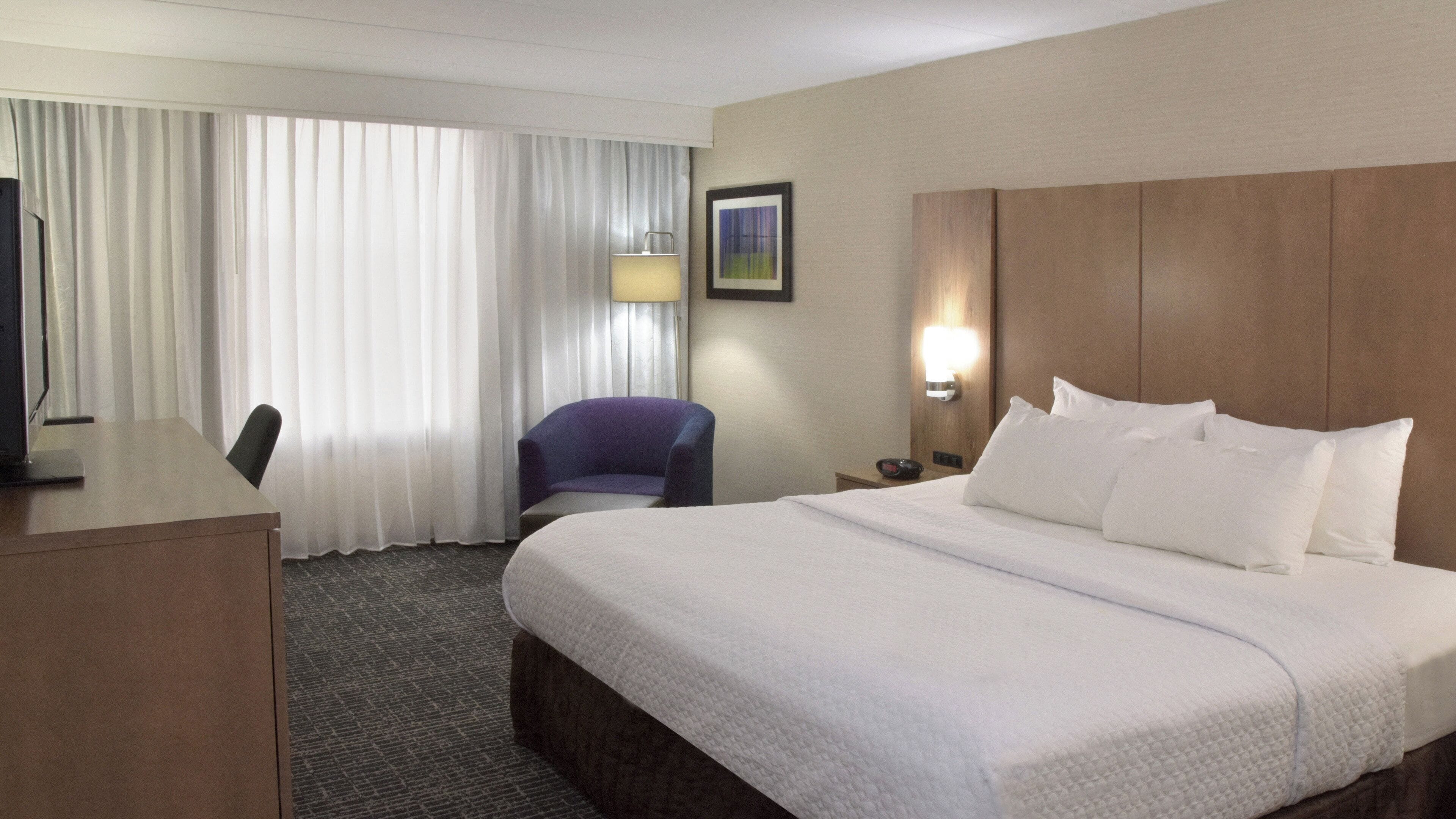 A hotel room with a bed, a desk, a chair, a lamp, and a nightstand.