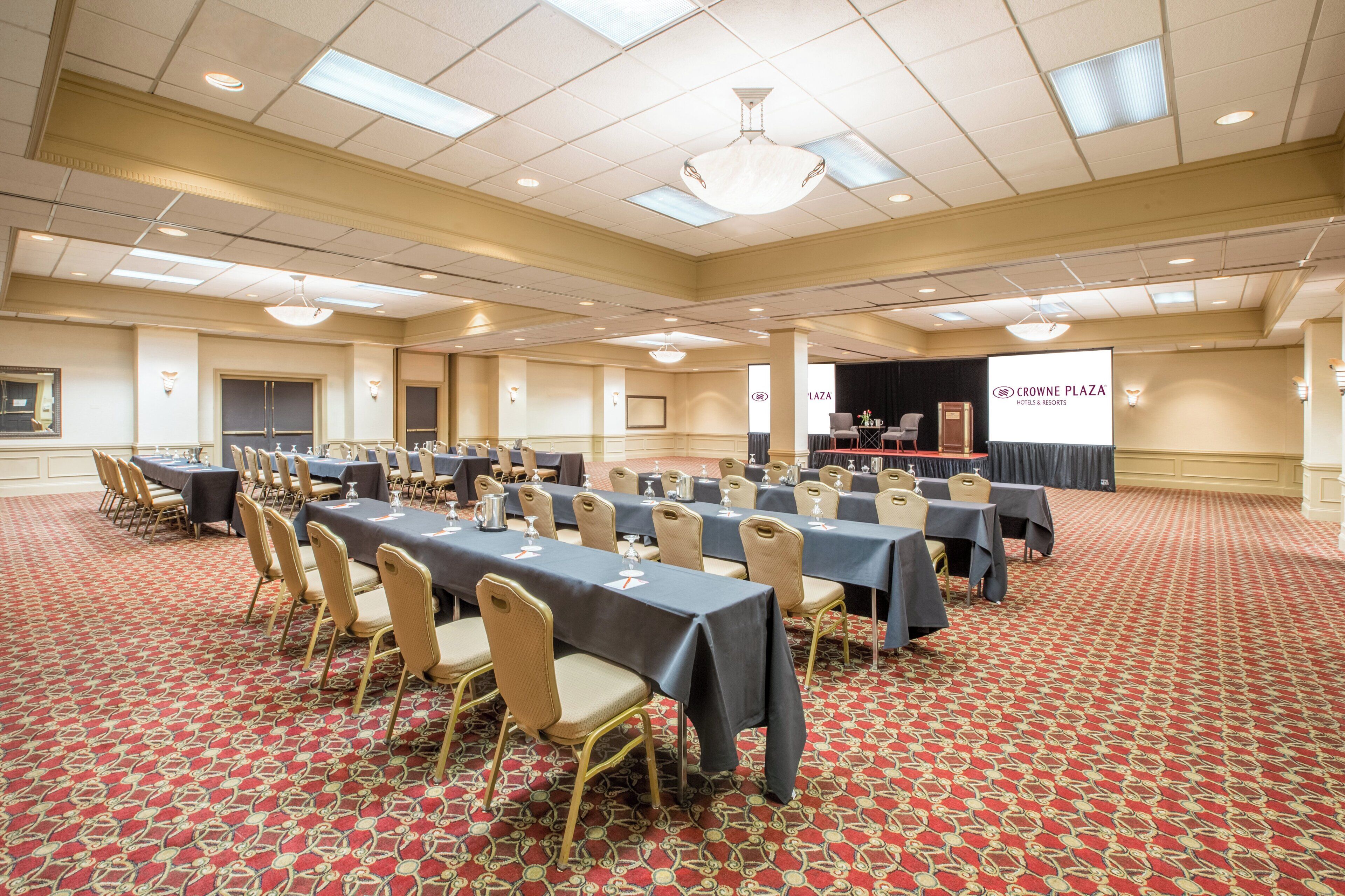 A conference room with multiple tables and chairs, a stage with a podium, and a projection screen displaying the Crowne Plaza Hotels & Resorts logo.