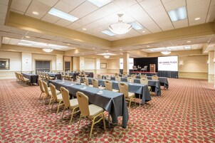 Un salón de conferencias con varias mesas y sillas, un escenario con un atril y una pantalla que muestra el logotipo de Crowne Plaza Hotels & Resorts.