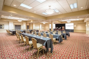 Un salón de conferencias con varias mesas y sillas, un escenario con un atril y una pantalla que muestra el logotipo de Crowne Plaza Hotels & Resorts.