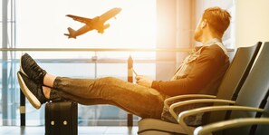 A person sitting on a chair with a suitcase, holding a mobile device, and an airplane is visible through a window. - Crowne Plaza Cleveland Airport by IHG (Middleburg Heights)
