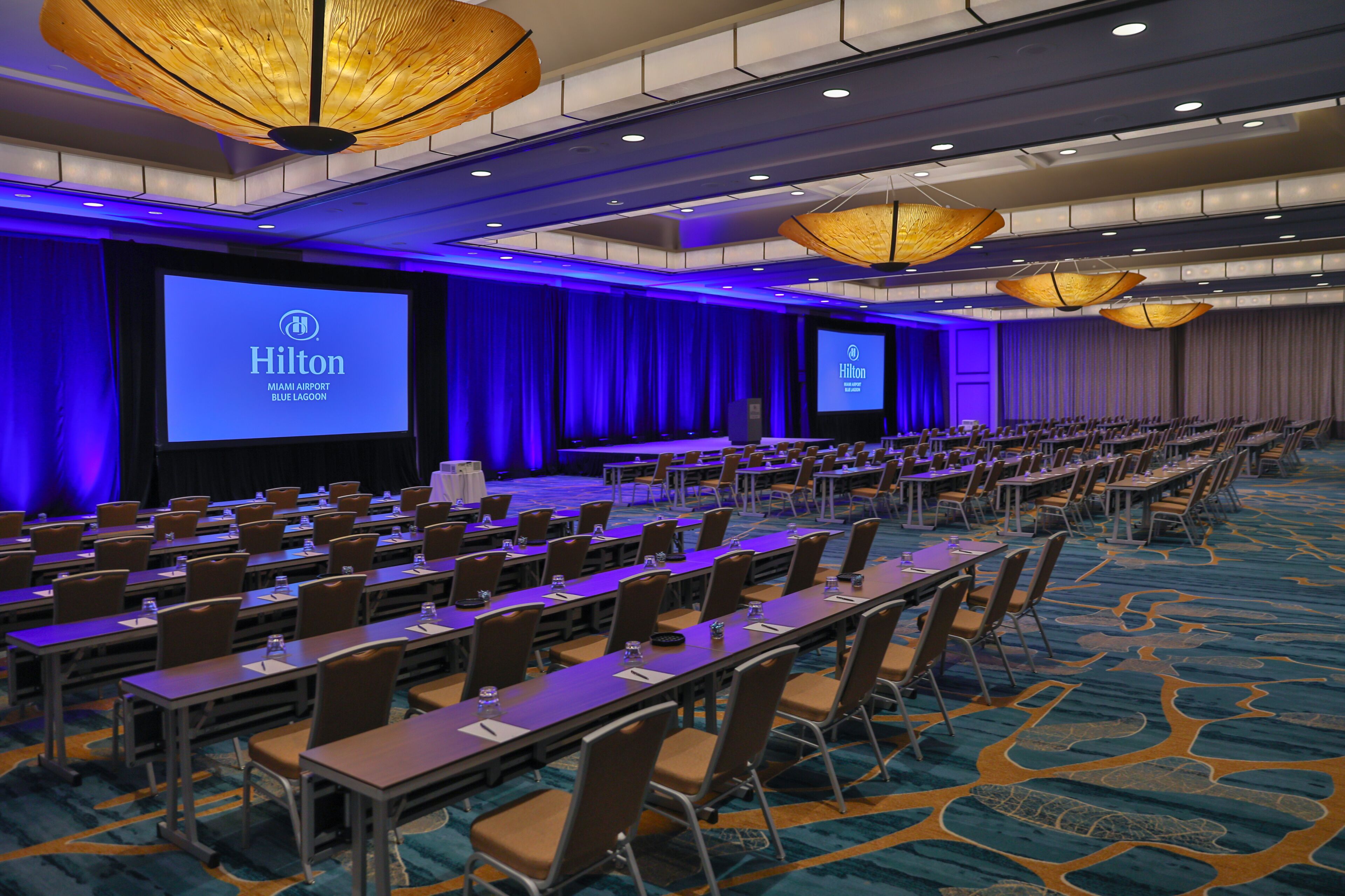 Une salle de conférence avec des rangées de tables et de chaises, de grands écrans affichant le logo de Hilton et un plafond éclairé en bleu.