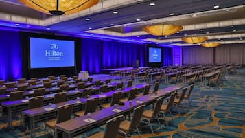 Une salle de conférence avec des rangées de tables et de chaises, de grands écrans affichant le logo de Hilton et un plafond éclairé en bleu.