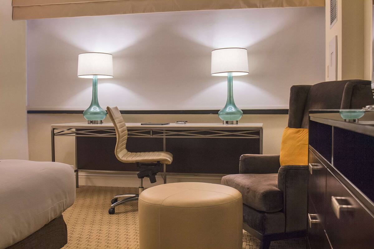 A hotel room with a bed, a desk, two lamps, a chair, and an ottoman.