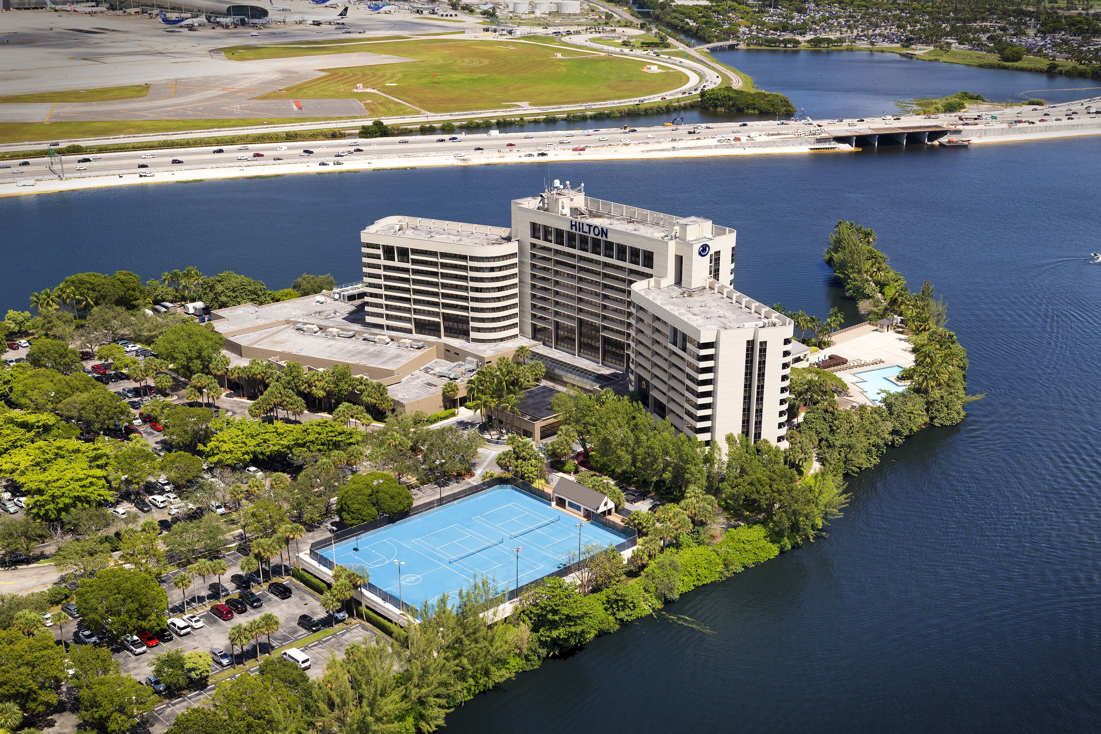 Un grand hôtel doté d’une piscine entourée de verdure, situé près d’une étendue d’eau et d’une autoroute.