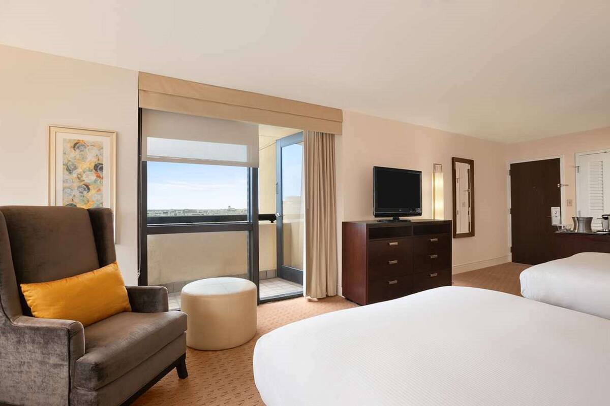 A hotel room with a large bed, a sofa with a cushion, a television, a desk with a lamp, and a sliding glass door leading to a balcony.