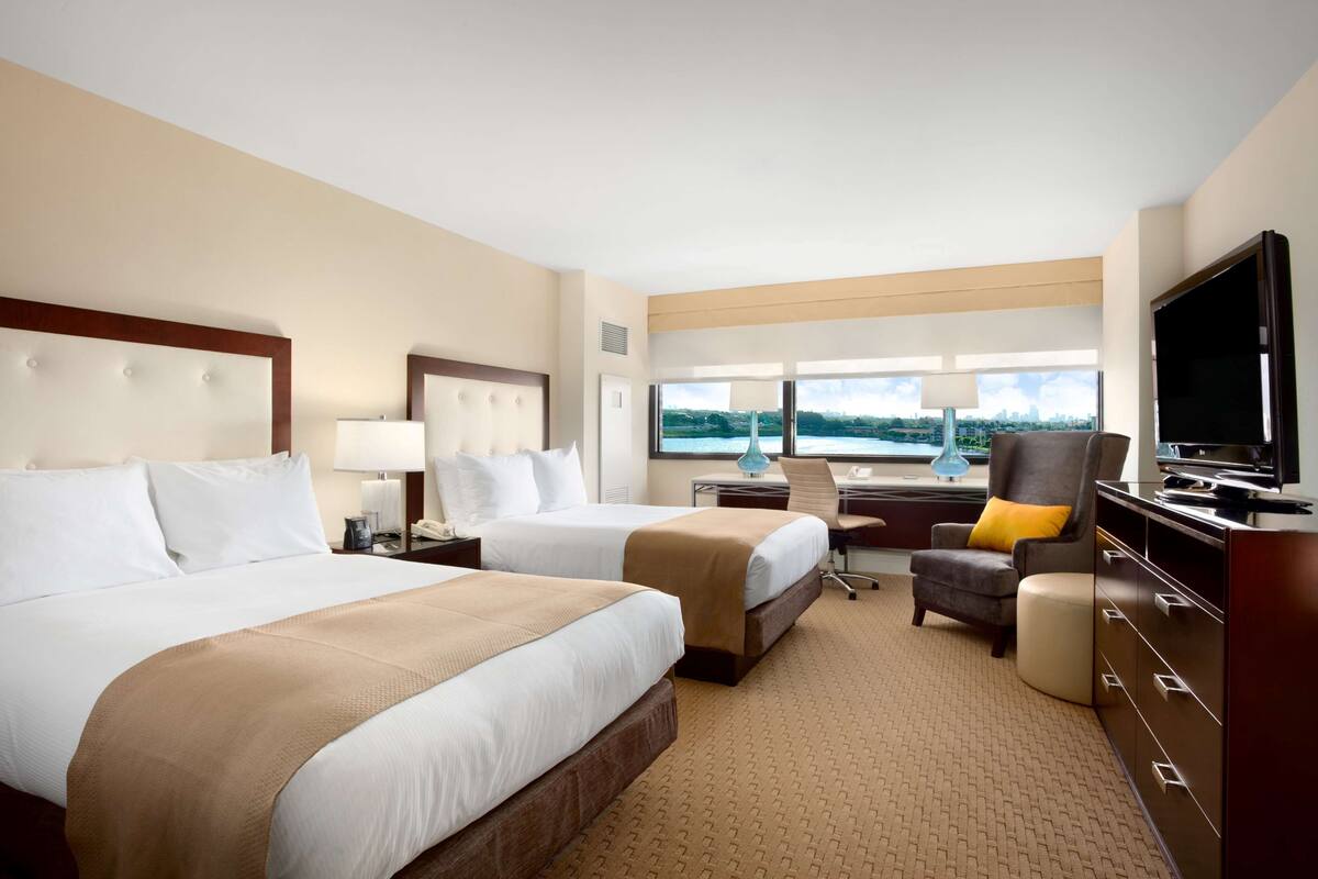 A hotel room with two beds, a desk, a chair, a TV, and a window with a city view.