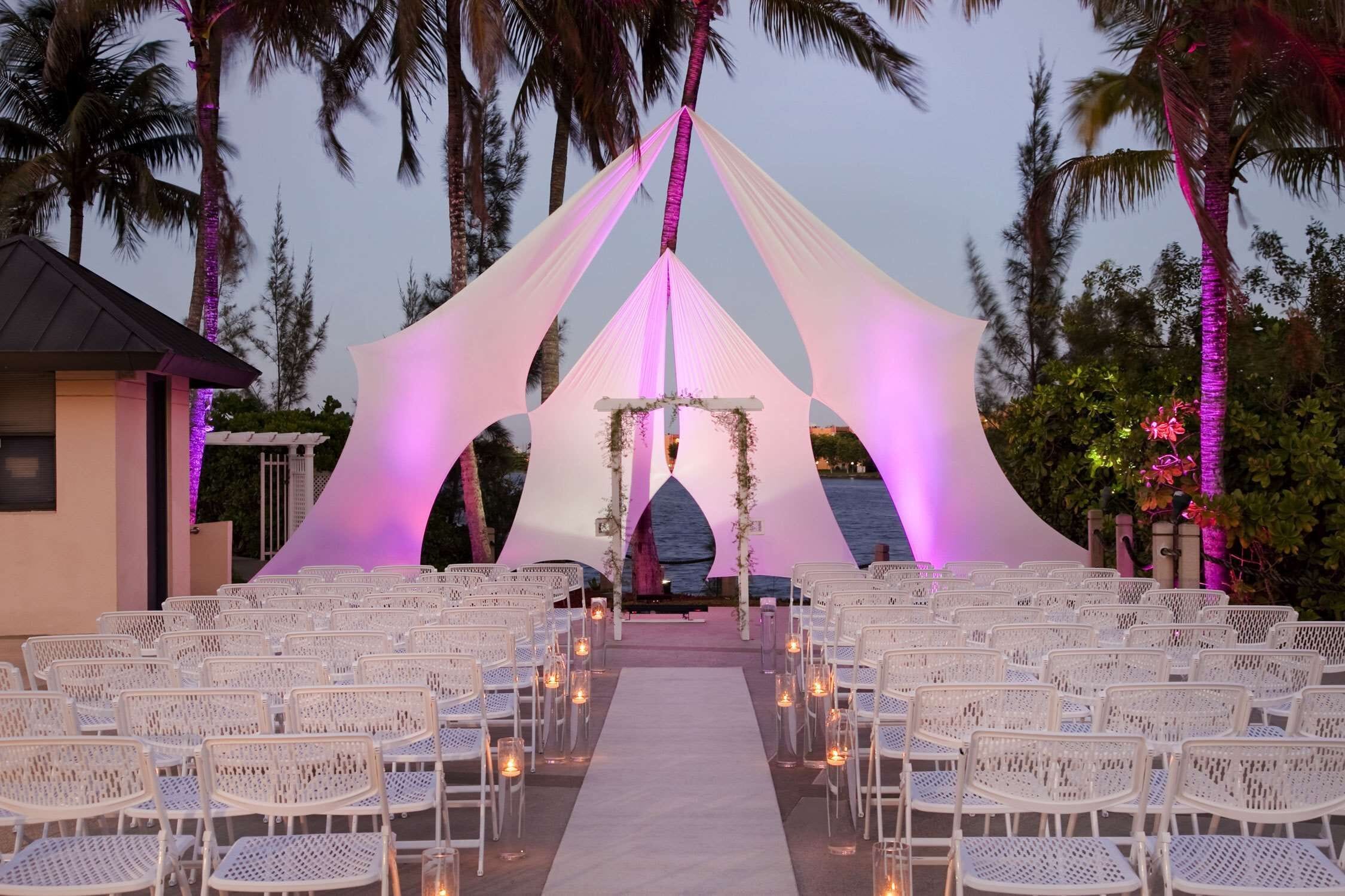 An outdoor wedding venue with white chairs arranged in rows, a central aisle, and a backdrop with draped fabric and floral decorations.