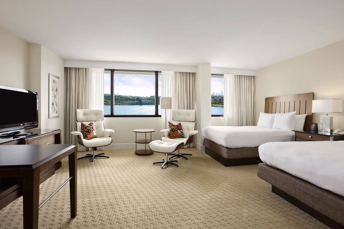 A hotel room with a bed, a desk with a television, two chairs, a small table, and a view of a lake.