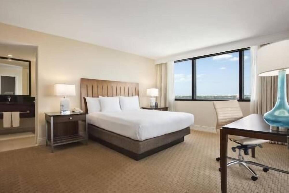 A hotel room with a bed, bedside tables, a desk, a chair, a large window, and a view of the sky.
