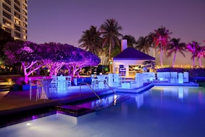 Un espace piscine aménagé pour la nuit, avec un fauteuil de relaxation et un parasol, entouré de palmiers et d’un bâtiment dont les fenêtres sont illuminées.