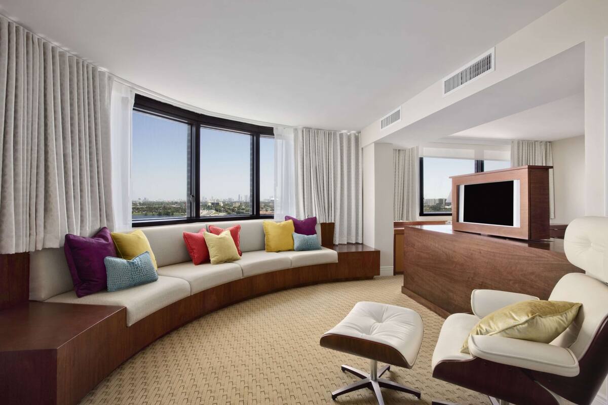 A modern hotel room with a curved sofa, a flat-screen TV, a white armchair, and a view of the city through large windows.